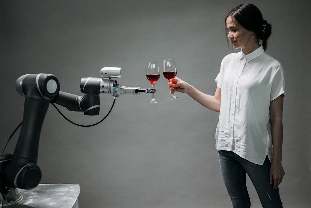 A robotic arm holds up a glass of red wine to cheers a young woman with brown hair in a white shirt, who also holds a glass of red wine.