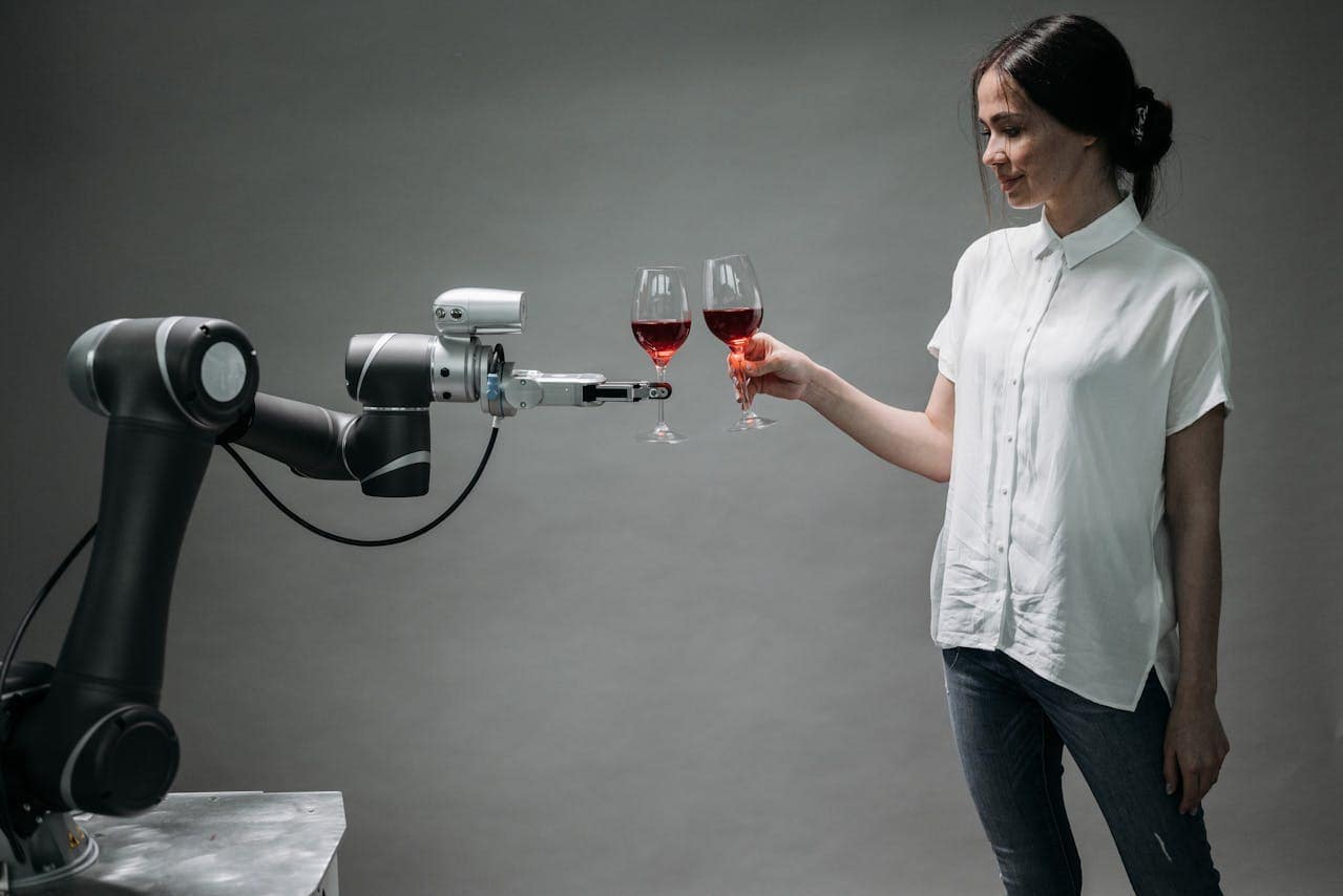 A robotic arm holds up a glass of red wine to cheers a young woman with brown hair in a white shirt, who also holds a glass of red wine.