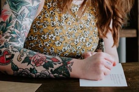 A red haired woman with arm tatoo's writes with a pen at a desk by a book shelf.