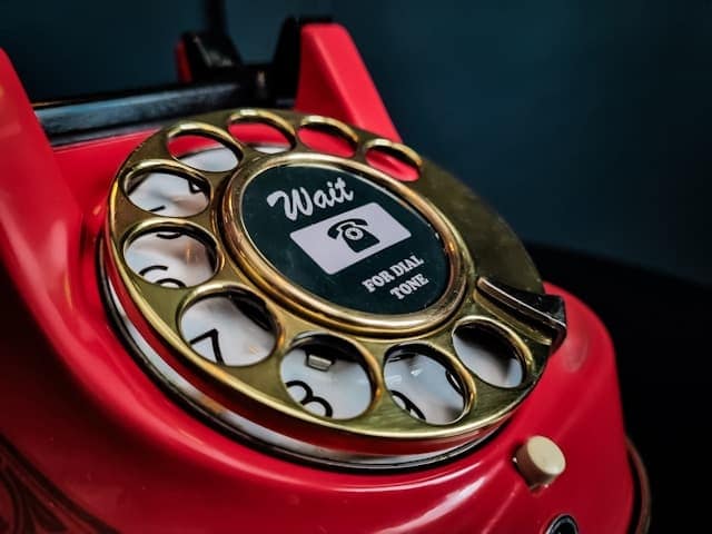 A close up of a red rotary dial phone with a gold dialler, saying "wait for the tone".